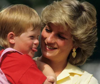 A young boy with blond hair playfully sticks out his tongue, held by a smiling woman with short blond hair and gold earrings.