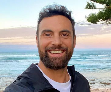 Smiling man with short hair and a beard stands near a beach at sunset, wearing a dark jacket and a white shirt.
