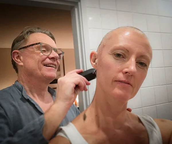 Man shaving a woman's head in a bathroom, both appear calm and supportive.