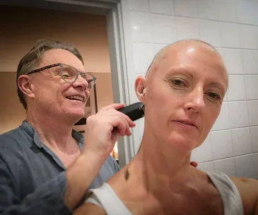 Man shaving a woman's head in a bathroom, both appear calm and supportive.