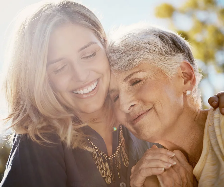 Elderly woman and younger woman smiling and embracing warmly in sunlight.