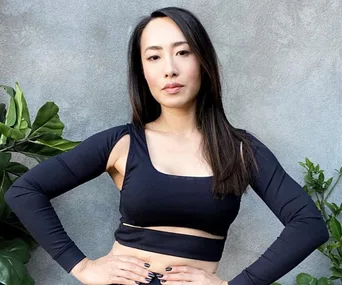 Woman in a stylish black outfit posing confidently against a gray wall with plants.
