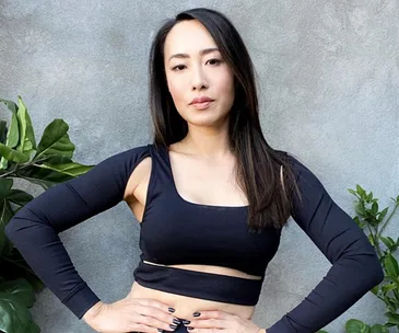 Woman in a stylish black outfit posing confidently against a gray wall with plants.