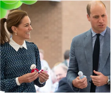Prince and Princess holding knitted toys, smiling during a hospital visit with green balloons in the background.