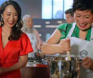 Two chefs in a kitchen; one in a red outfit, smiling, and the other wearing a green apron, stirring a bowl.