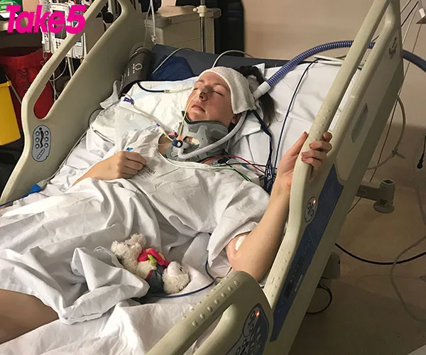 A person lies unconscious in a hospital bed, connected to medical equipment and wearing a neck brace.