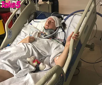 A person lies unconscious in a hospital bed, connected to medical equipment and wearing a neck brace.