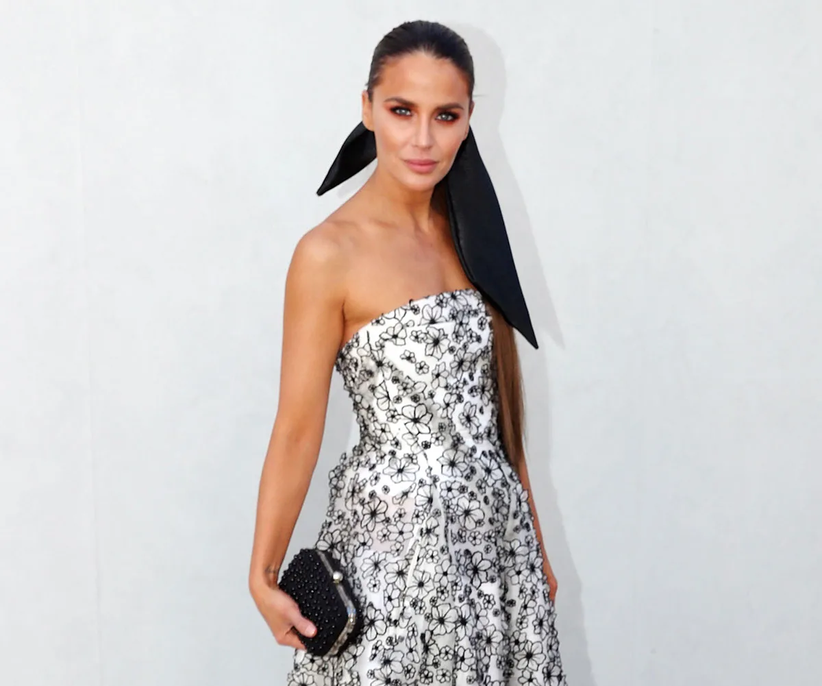 A person in a strapless floral dress, holding a clutch, with a backdrop of a plain light wall.