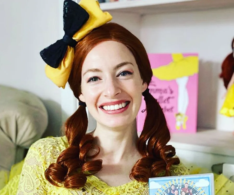 Person smiling in a yellow top with a large yellow and black bow in their red hair, holding a colorful book.