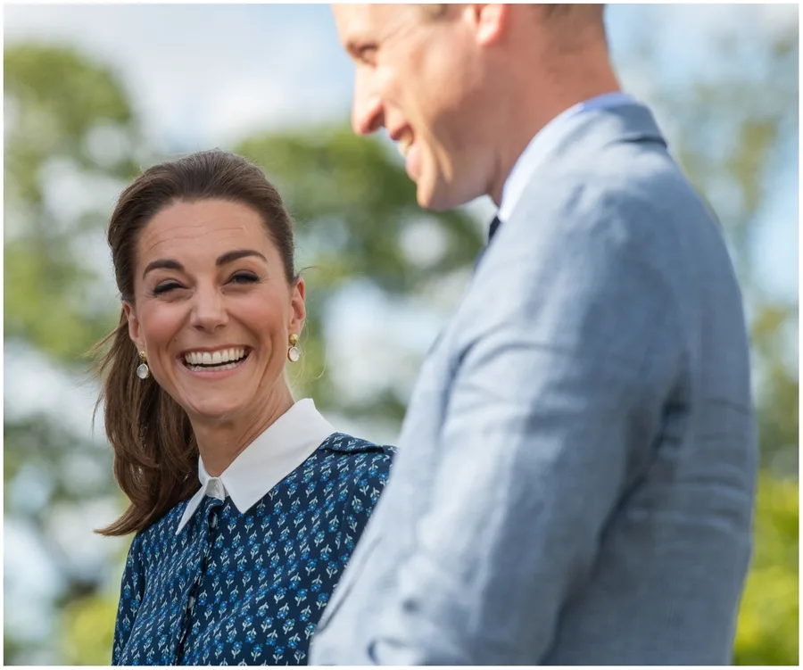 A smiling woman in a blue patterned outfit stands beside a man in a light blue suit, outdoors.