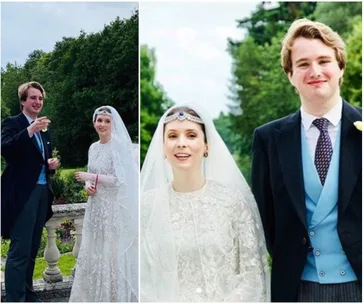 Bride in ornate white gown and veil stands with groom in formal suit amid lush greenery, both smiling.