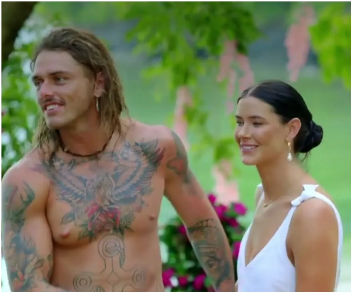 A shirtless man with tattoos stands next to a woman in a white dress, both smiling in a garden setting.