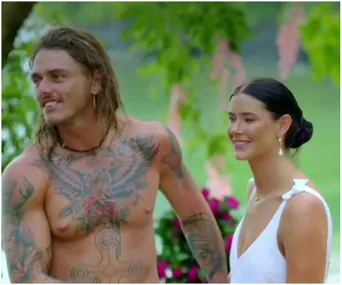 A shirtless man with tattoos stands next to a woman in a white dress, both smiling in a garden setting.
