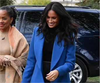 A smiling woman in a blue coat walks outdoors with another woman, in front of a parked car.
