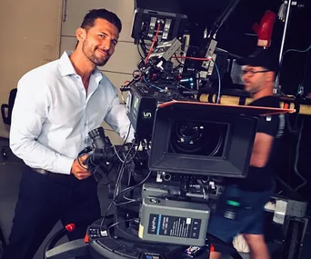 Man in a white shirt stands next to filming equipment, smiling at the camera.