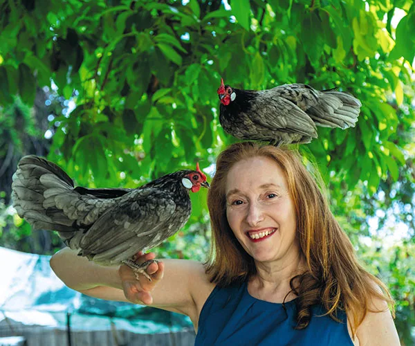 A woman smiles with one chicken perched on her head and another on her hand under a leafy tree.