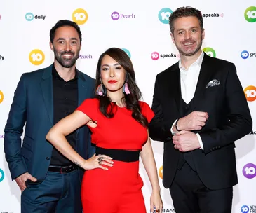Three people posing on a red carpet, wearing formal attire, with logo backdrop.