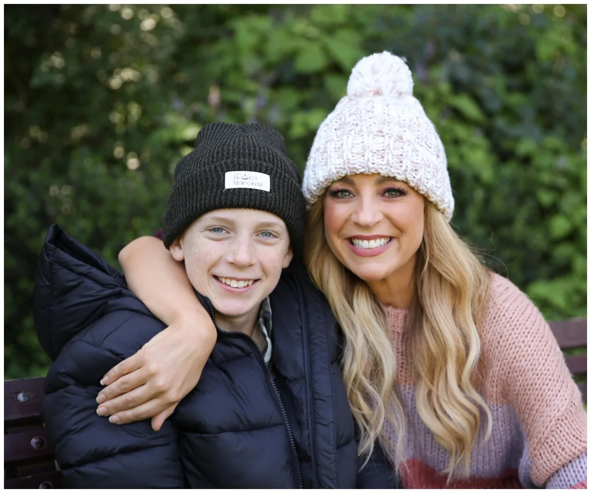A woman and a boy wearing beanies, smiling on a bench, surrounded by greenery.