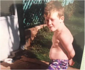 Young boy with short hair standing shirtless outdoors, wearing colorful shorts, in a garden with grass and a wooden fence.