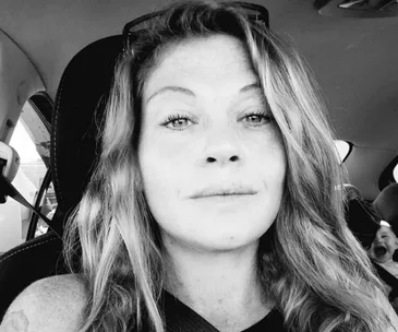 Person with long hair sitting in a car, looking at the camera, in black and white.