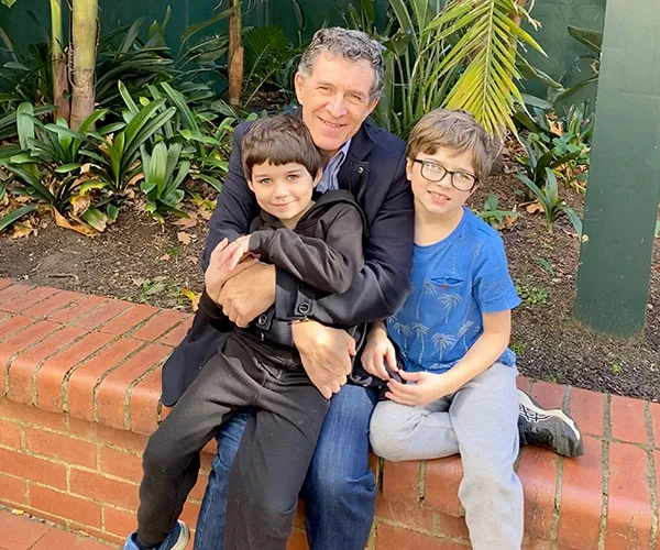 Man sitting on a brick ledge hugs two smiling boys in a garden setting with lush greenery.