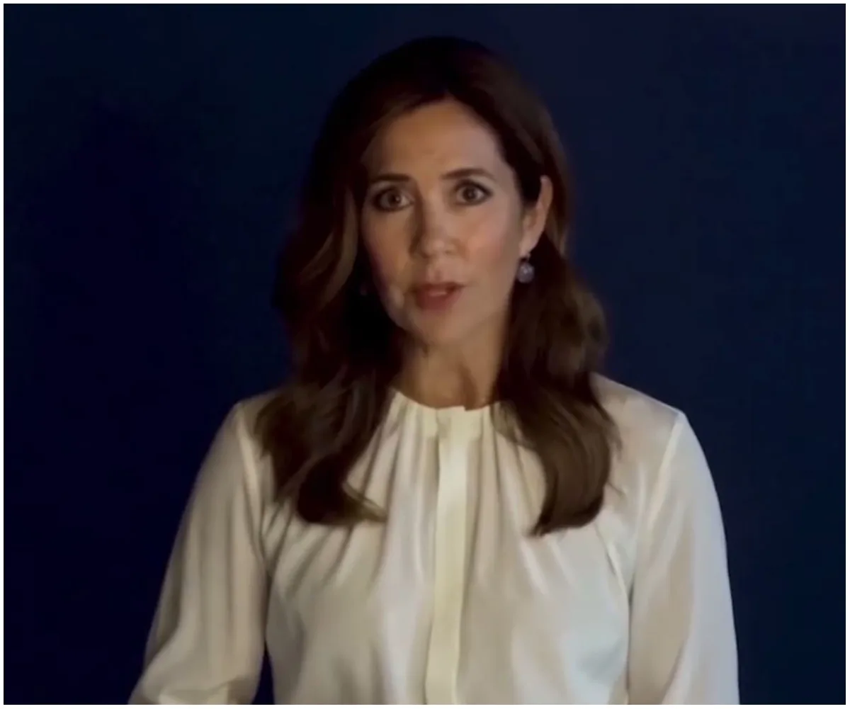 Woman in white blouse speaking, against a dark blue background.
