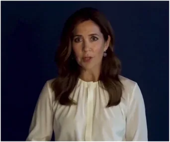 Woman in white blouse speaking, against a dark blue background.