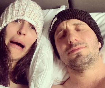 Couple in bed wearing beanies; one making a funny face, the other relaxed. Cozy moment captured on camera.