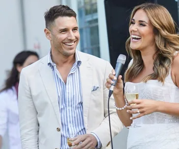 Man and woman smiling at an outdoor event; the woman holds a microphone and a glass.