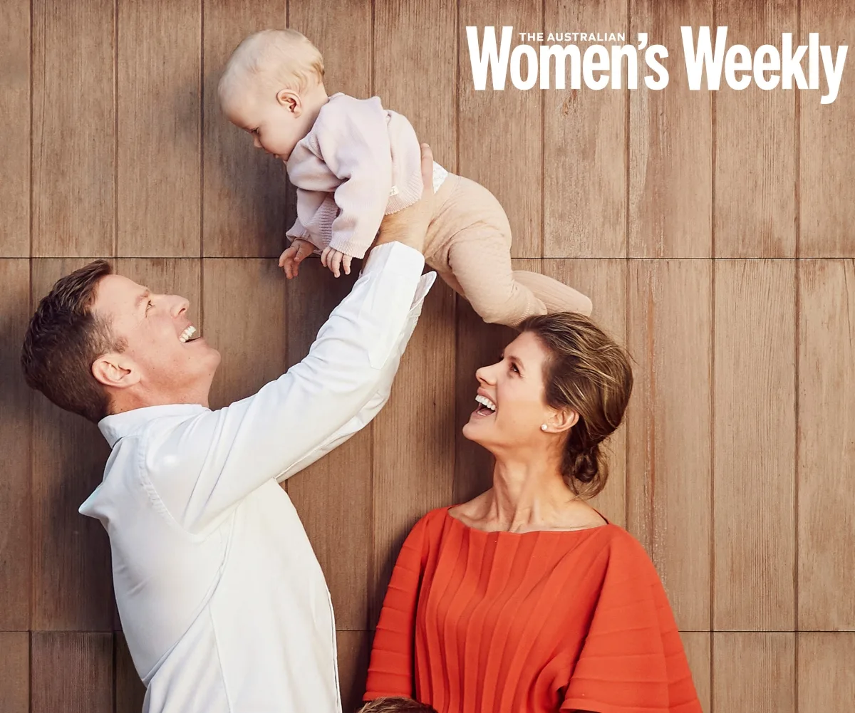 Man in white shirt lifts baby, woman in orange top smiles, wood panel background.