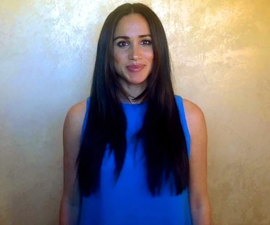 Individual in a blue sleeveless top stands against a light textured wall, smiling at the camera.