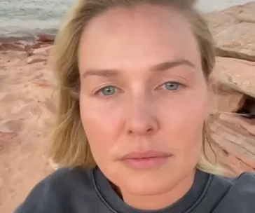 A person with blonde hair and a serene expression, outdoors with sandy rocks in the background.