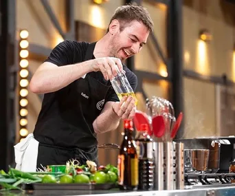Chef preparing a dish in a kitchen, wearing a black apron and holding a jar of oil with various cooking utensils nearby.
