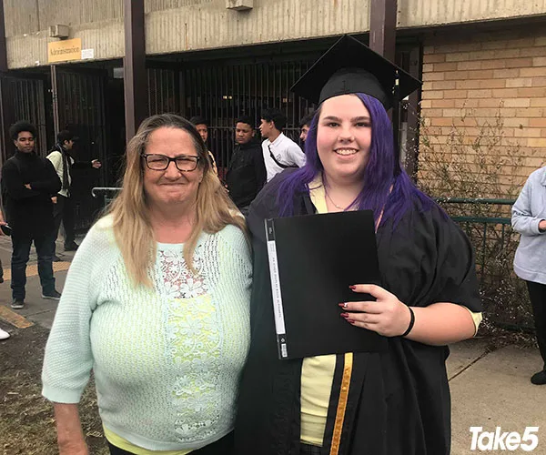 "Young woman in graduation gown holds diploma, posing with an older woman outdoors."