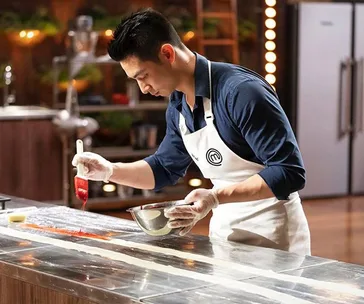Chef in a MasterChef apron painting on a metal counter with focus and precision.