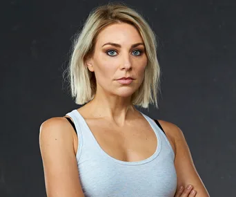 A woman with short blonde hair in a sleeveless top stares seriously at the camera against a dark background.