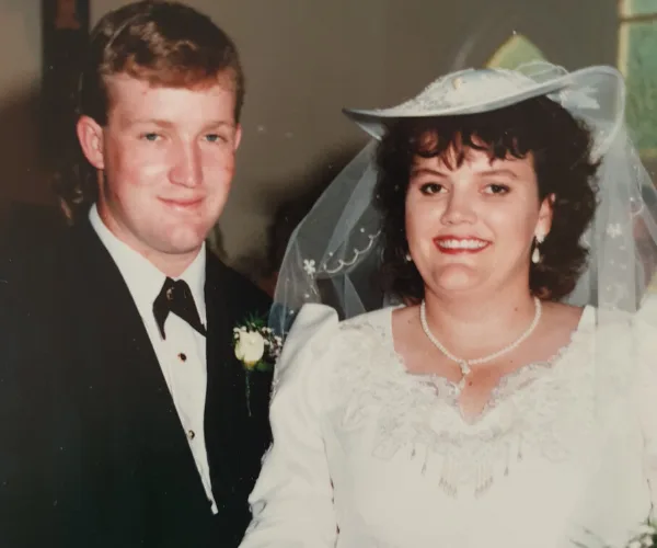 Wedding photo of a smiling couple; the bride in a white dress and veil, the groom in a black suit and tie.