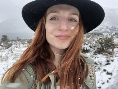 Young woman with red hair in a snow-covered landscape, wearing a black hat, green jacket, and smiling gently.