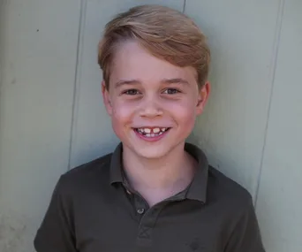 Young boy smiling, wearing a dark polo shirt, standing against a light-colored wall.