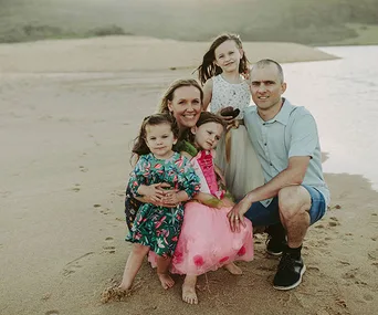 A family of five poses on a sandy beach, with three young girls in colorful dresses and the parents smiling at the camera.