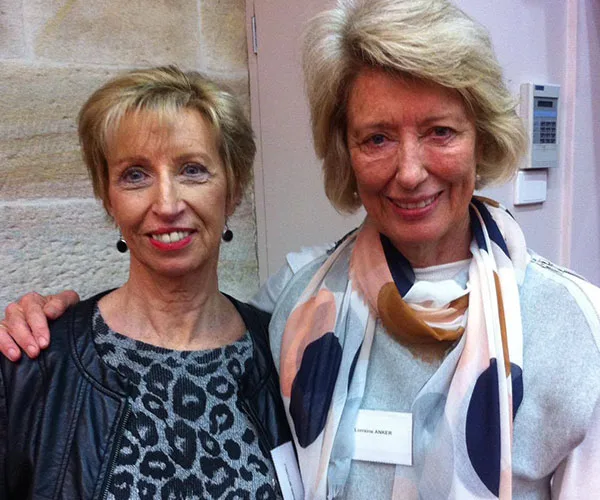 Two women smiling, standing side by side indoors; one wears a patterned top and leather jacket, the other a scarf and jacket.
