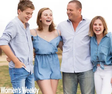 A family smiling and laughing outdoors, wearing casual blue outfits, appearing happy and relaxed.