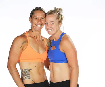 Two women smiling, wearing athletic gear; one in an orange top, the other in blue, showcasing tattoos.