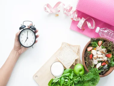Clock, healthy meal, water bottle, pink yoga mat, and measuring tape on white background, symbolizing weight loss and fitness.