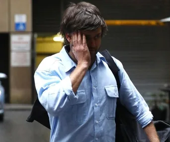 Man in a blue shirt covers his face with one hand while walking in an urban setting, holding a bag.