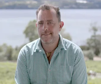 A man in a checked shirt smiling outdoors with a scenic lake and trees in the background.