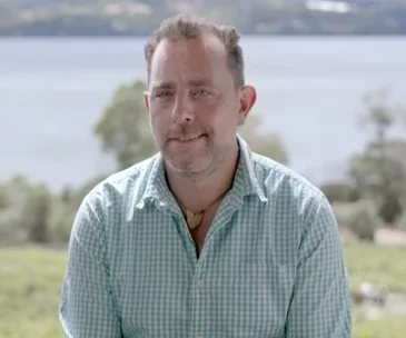A man in a checked shirt smiling outdoors with a scenic lake and trees in the background.