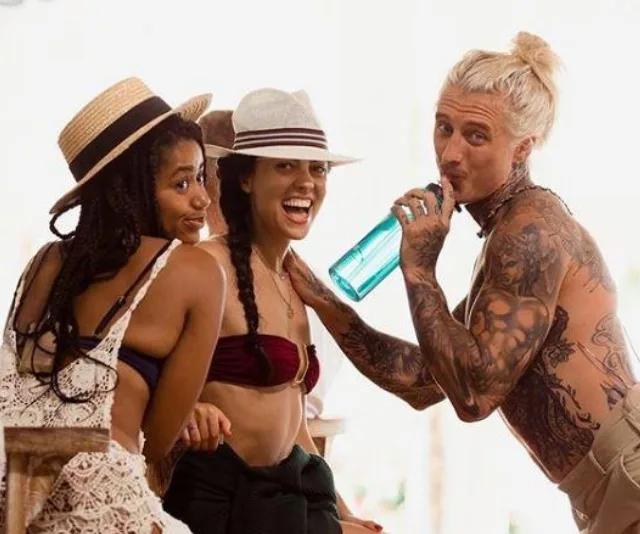 Three people smiling, wearing hats, with one drinking from a blue bottle on a sunny day.