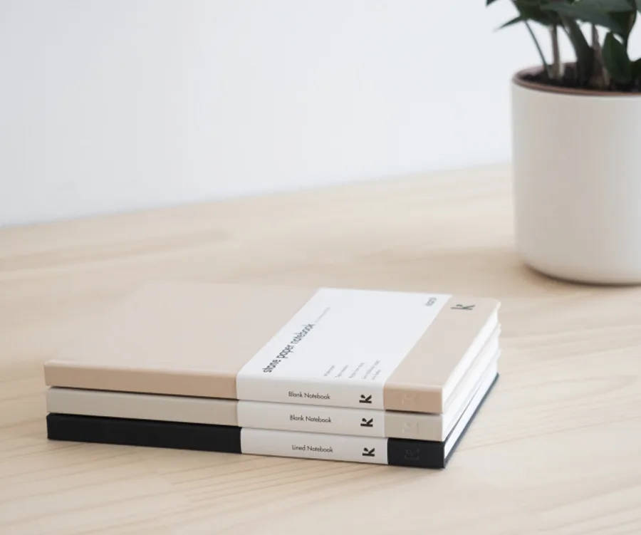 Stack of three notebooks on a wooden table with a potted plant in the background.