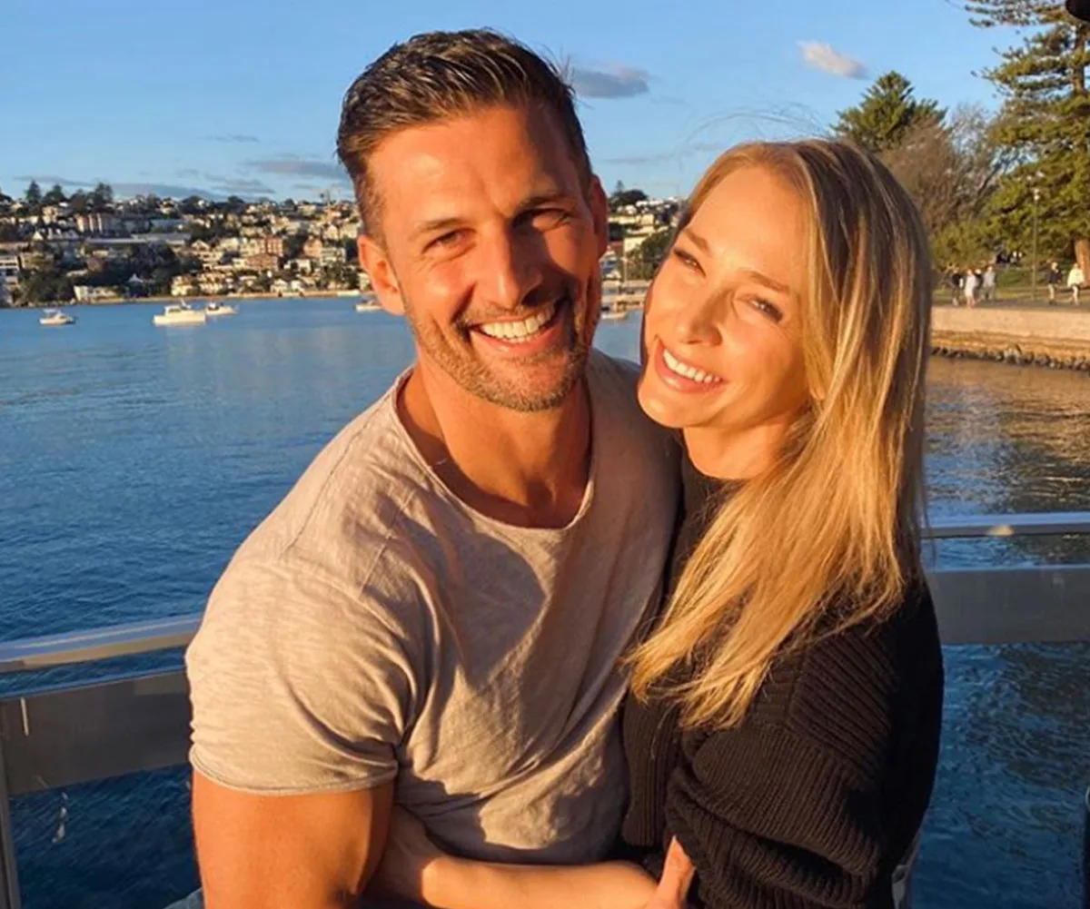 A smiling couple embraces on a sunny waterfront with boats and buildings in the background.